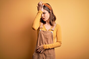 Young beautiful blonde girl wearing overall standing over yellow isolated background surprised with hand on head for mistake, remember error. Forgot, bad memory concept.