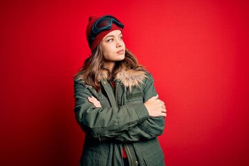 Fototapeta premium Young blonde girl wearing ski glasses and winter coat for ski weather over red background looking to the side with arms crossed convinced and confident