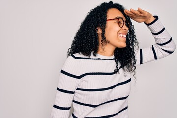 Young african american woman wearing striped sweater and glasses over white background very happy and smiling looking far away with hand over head. Searching concept.