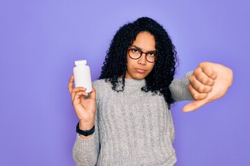 Young african american curly woman holding pills standing over isolated purple background with angry face, negative sign showing dislike with thumbs down, rejection concept