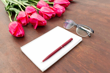 Pink tulips on the wooden table with empty white note, pen and glasses