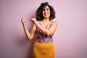 Young beautiful arab woman on vacation wearing swimsuit and sunglasses over pink background Pointing to the back behind with hand and thumbs up, smiling confident