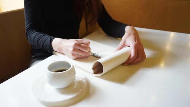 a Hand of a girl draws in sketchbook with a pencil in a cafe, a cup of coffee on the table, long Travelling shot