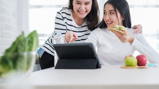 Happy Asian business friend having conversation in office.Two young business women sitting at table at home.  Teamwork, business meeting. Freelancers .eating breakfast sandwich.