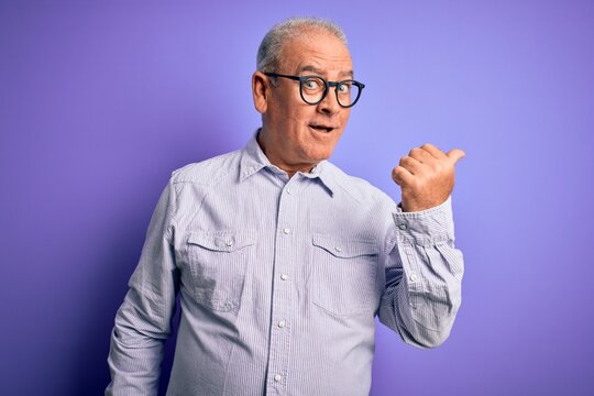 Middle age handsome hoary man wearing striped shirt and glasses over purple background smiling with happy face looking and pointing to the side with thumb up.
