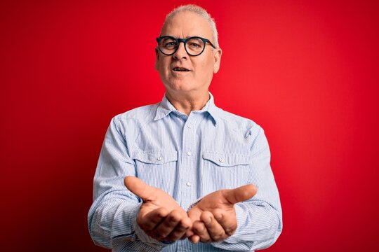 Middle age handsome hoary man wearing casual striped shirt and glasses over red background Smiling with hands palms together receiving or giving gesture. Hold and protection
