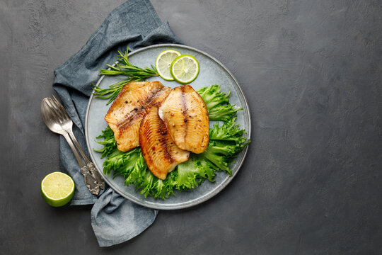 Grilled fish sea bass fillet with green lettuce in a plate on a black stone background. Above view.