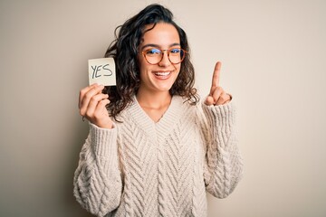 Young beautiful woman with curly hair holding reminder paper with yes word message surprised with...