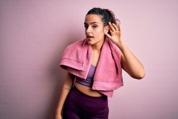 Young beautiful sportswoman with curly hair doing sport using towel over pink background smiling with hand over ear listening an hearing to rumor or gossip. Deafness concept.