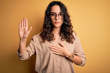 Beautiful woman with curly hair wearing striped shirt and glasses over yellow background Swearing...