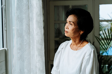 Close up of pensive old woman looking out of window feeling lonely. 70s Asian elderly female...