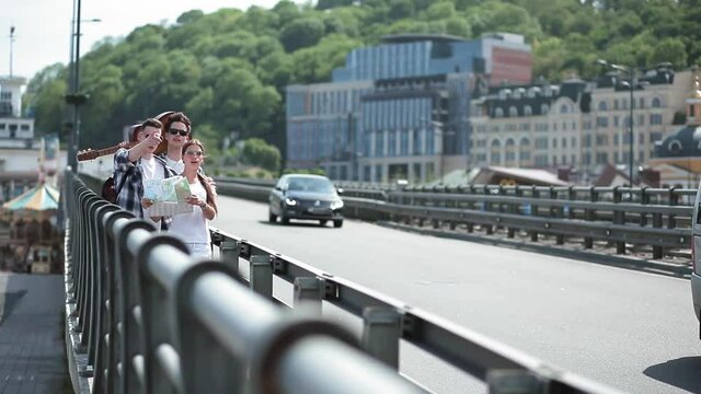 Friends walking on viaduct with map and guitar on shoulder and cars passing by. Three friends are walking at urban city enjoying time together. Friends concept. August 2018. Kiev, Ukraine. Prores 422.