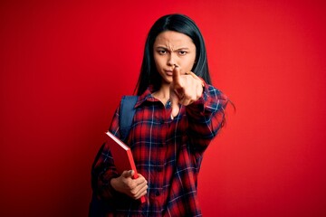 Naklejka premium Young beautiful chinese student woman holding book standing over isolated pink background pointing with finger to the camera and to you, hand sign, positive and confident gesture from the front