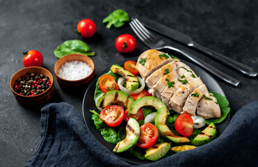 
Grilled chicken breast and grilled avocado salad with cherry tomatoes, spinach, in a black plate on a stone background
