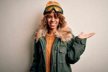 Young african american skier woman with curly hair wearing snow sportswear and ski goggles smiling cheerful presenting and pointing with palm of hand looking at the camera.