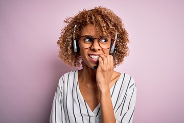 African american curly call center agent woman working using headset over pink background looking stressed and nervous with hands on mouth biting nails. Anxiety problem.