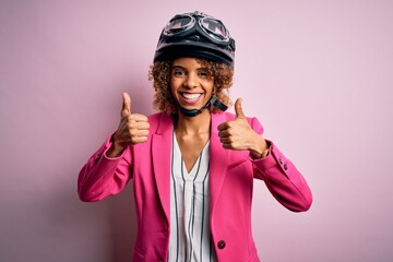 African american motorcyclist woman with curly hair wearing moto helmet over pink background success sign doing positive gesture with hand, thumbs up smiling and happy. Cheerful expression and winner