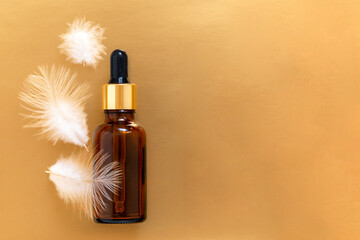 Fototapeta premium Oil bottle with pipette on the golden background with white feathers and copy space. Skin care concept