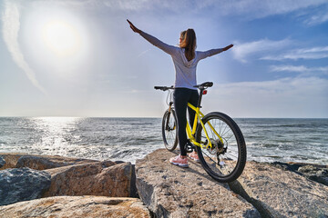 Free woman with bicycle stands on the stones on the seashore and raises hands to the sky as a sign...