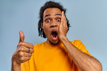 Portrait of emotional shocked dark skinned young guy opens mouth from wonder, stares at camera, raises thumbs up, fascinated by something awesome, demonstrates okay gesture on blue background