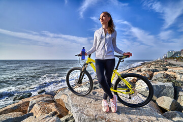 Fitness woman with yellow bicycle stands on the stones on the seashore in a clear sunny day against the blue sky. Athletic healthy people with active and sporty lifestyle