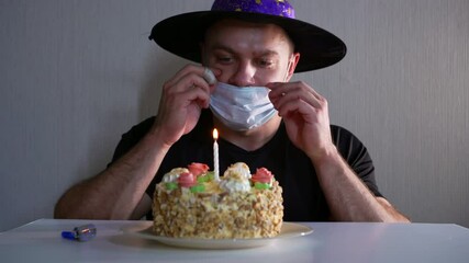 Portrait of a man who sits at home on self-isolation and alone celebrates his birthday with a cake. He has a hat on his head and is trying to blow out a candle through a mask on his face. - Powered by Adobe