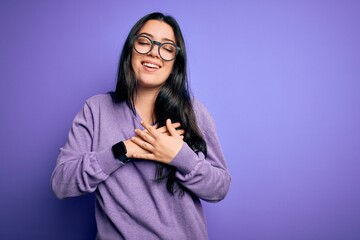 Young brunette woman wearing glasses over purple isolated background smiling with hands on chest with closed eyes and grateful gesture on face. Health concept.