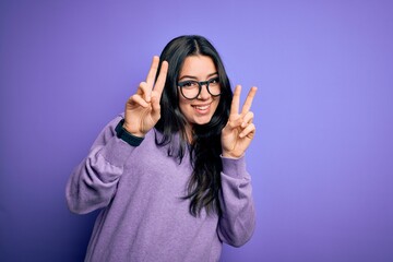 Young brunette woman wearing glasses over purple isolated background smiling looking to the camera showing fingers doing victory sign. Number two.