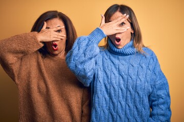 Middle age beautiful couple of sisters wearing casual sweater over isolated yellow background peeking in shock covering face and eyes with hand, looking through fingers with embarrassed expression.