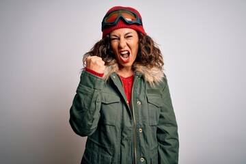 Middle age skier woman wearing snow sportswear and ski goggles over white background angry and mad raising fist frustrated and furious while shouting with anger. Rage and aggressive concept.