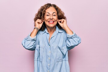 Middle age beautiful woman wearing casual denim shirt standing over pink background Smiling pulling ears with fingers, funny gesture. Audition problem