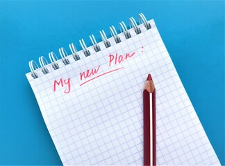 In a spiral notebook in a cage in red pencil is written "My new plan" and underlined. Red pencil on a notebook, all on a bright blue background