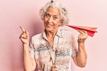 Senior grey-haired woman holding paper airplane smiling happy pointing with hand and finger to the side