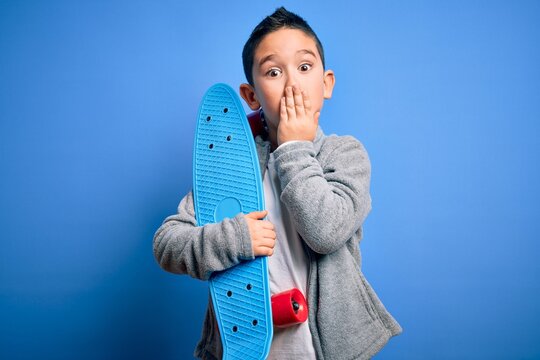Young little boy kid skateboarder holding modern skateboard over blue isolated background cover mouth with hand shocked with shame for mistake, expression of fear, scared in silence, secret concept