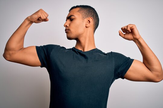 Young handsome african american man wearing casual t-shirt standing over white background showing arms muscles smiling proud. Fitness concept.