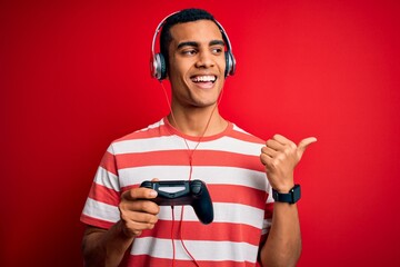 Handsome african american gamer man playing video game using jostick and headphones pointing and showing with thumb up to the side with happy face smiling