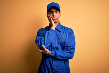 Young african american mechanic man wearing blue uniform and cap over yellow background thinking looking tired and bored with depression problems with crossed arms.