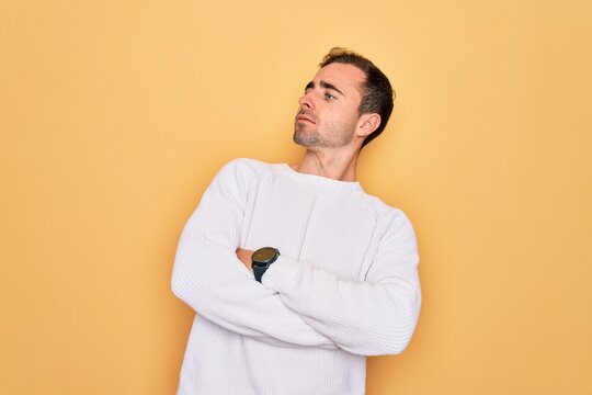 Young handsome man with blue eyes wearing casual sweater standing over yellow background looking to the side with arms crossed convinced and confident