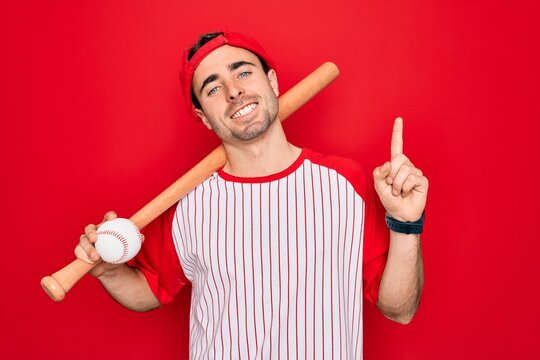 Young handsome sporty man with blue eyes playing baseball wearing cap holding bat and ball surprised with an idea or question pointing finger with happy face, number one