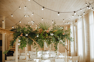 Restaurant, served table, rustic, huge compositions of flowers and tropical leaves on the table, plates, cutlery, crystal glasses, wooden chairs, garlands of light bulbs.