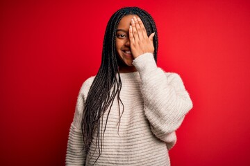 Young african american woman wearing casual winter sweater over red isolated background covering one eye with hand, confident smile on face and surprise emotion.