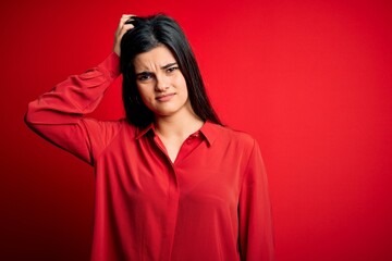 Young beautiful brunette woman wearing casual shirt standing over red background confuse and wonder...