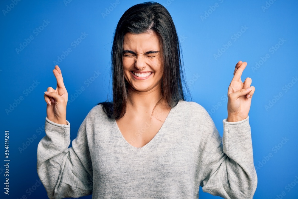 Wall mural Young beautiful brunette woman wearing casual sweater standing over blue background gesturing finger crossed smiling with hope and eyes closed. Luck and superstitious concept.