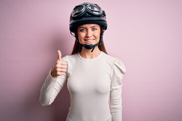 Young beautiful motorcyclist woman with blue eyes wearing moto helmet over pink background doing happy thumbs up gesture with hand. Approving expression looking at the camera showing success.