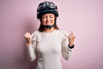 Young beautiful motorcyclist woman with blue eyes wearing moto helmet over pink background very happy and excited doing winner gesture with arms raised, smiling and screaming for success. Celebration