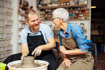 Senior woman spinning clay on a wheel with teacher at pottery class - obrazy, fototapety, plakaty