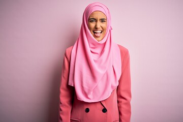 Fototapeta premium Young beautiful brunette businesswoman wearing pink muslim hijab and business jacket sticking tongue out happy with funny expression. Emotion concept.