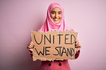 Young activist woman wearing pink muslim hijab holding banner with united we stand message with a...