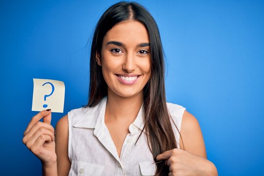 Young beautiful brunette woman holding paper with question mark symbol message with surprise face pointing finger to himself