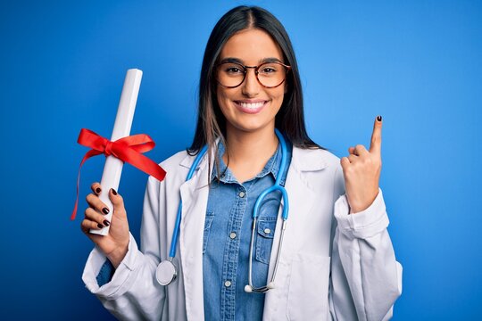 Young beautiful brunette doctor woman wearing glasses and coat holding diploma degree surprised with an idea or question pointing finger with happy face, number one